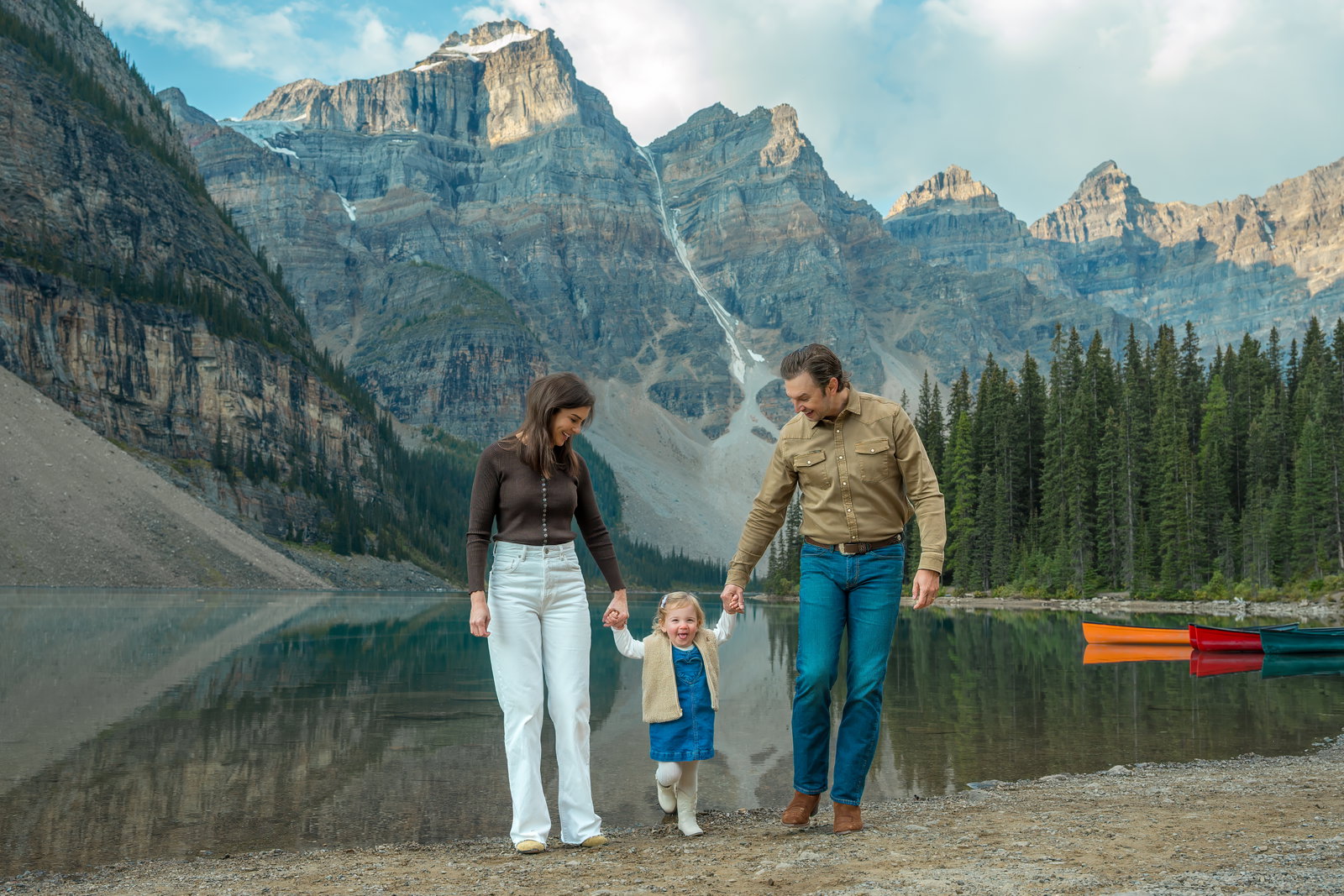 A family with a young child holding hands on the shore of Moraine Lake with turquoise water and mountain peaks in the background.