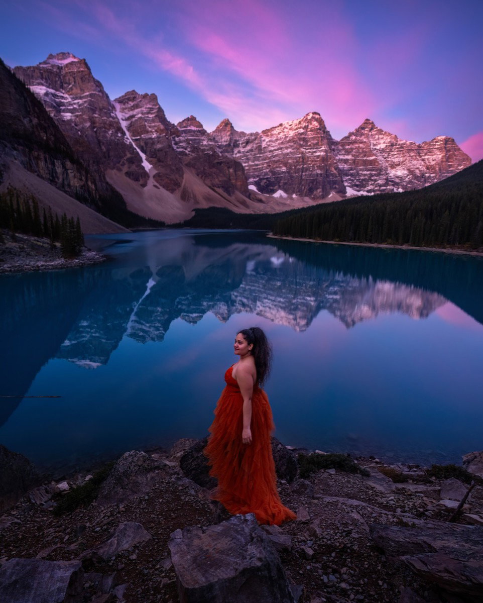 Woman in a red dress beside Moraine Lake under a pink sunrise sky
