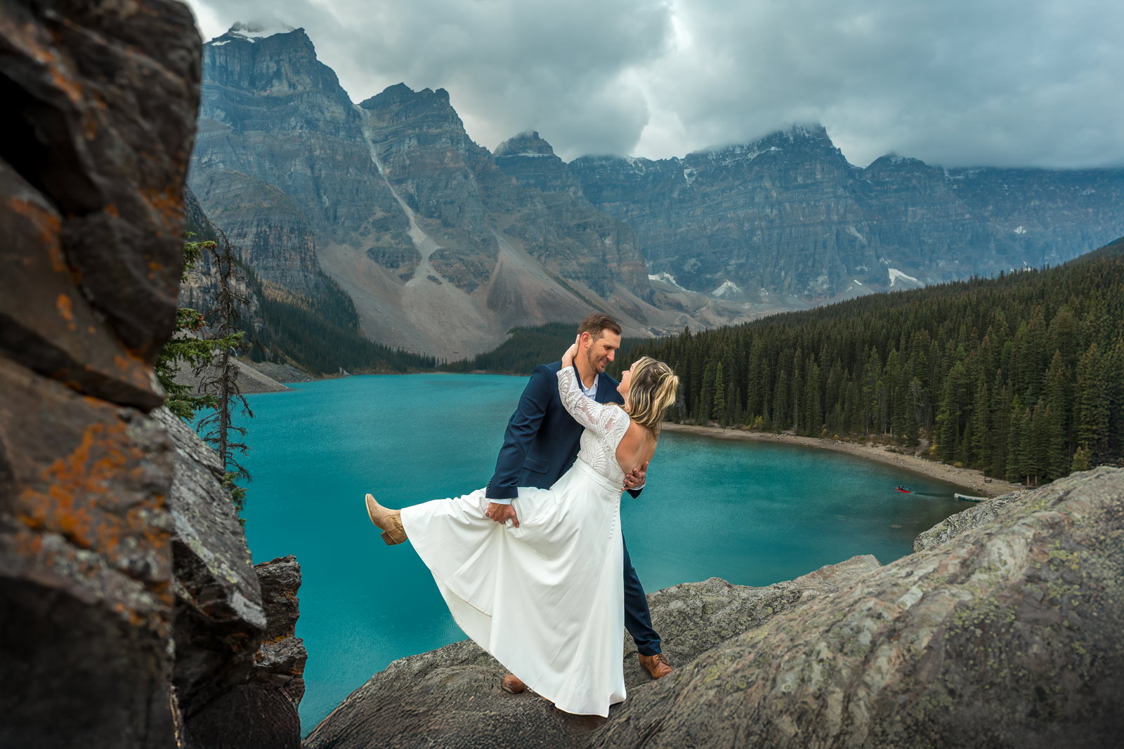 A groom dipping his bride in a white lace dress on rocks overlooking the turquoise waters of Moraine Lake.