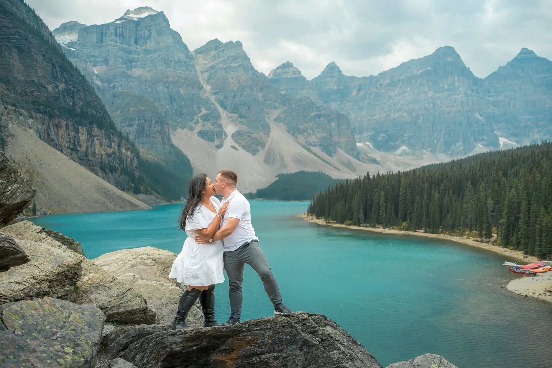 A couple in wedding attire standing hand in hand on a cliff edge, gazing at Moraine Lake and the surrounding mountains.