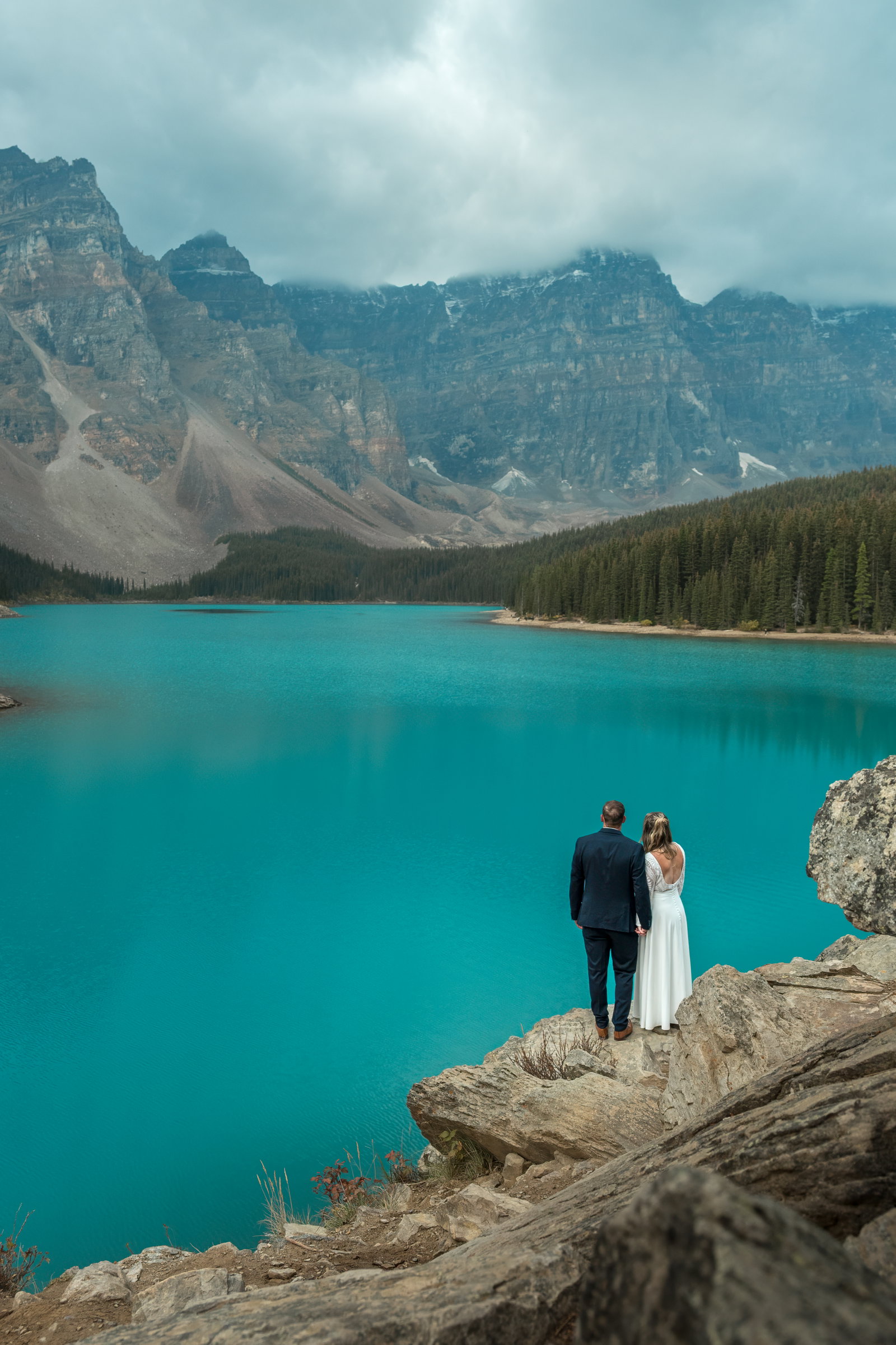 A couple in wedding attire sharing a kiss on a rocky overlook above Moraine Lake.