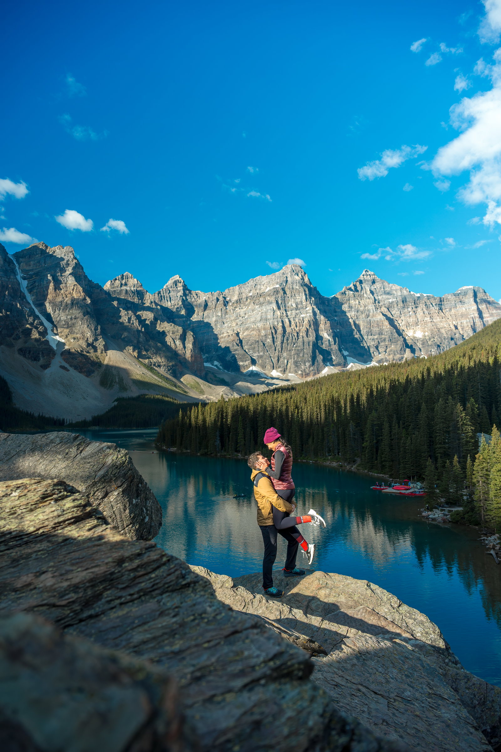 A joyful couple celebrating an engagement with a lift on a rocky outcrop overlooking Moraine Lake.
