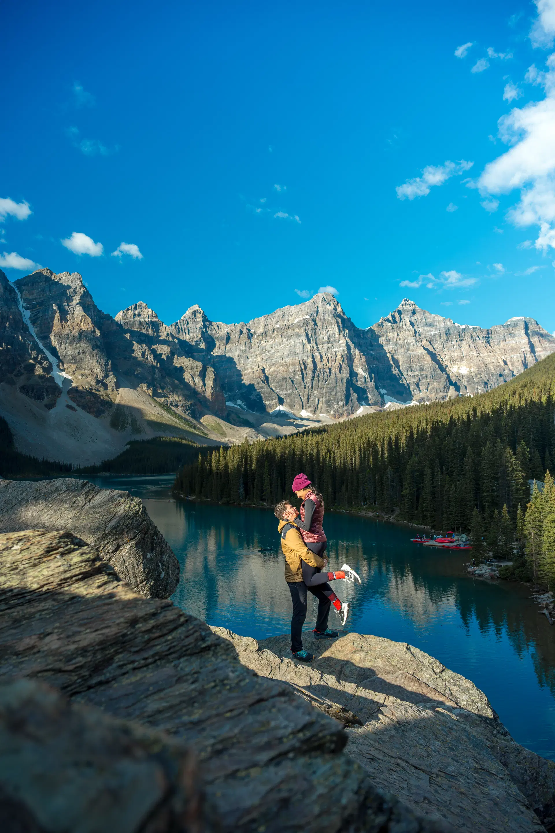 A joyful couple celebrating an engagement with a lift on a rocky outcrop overlooking Moraine Lake.