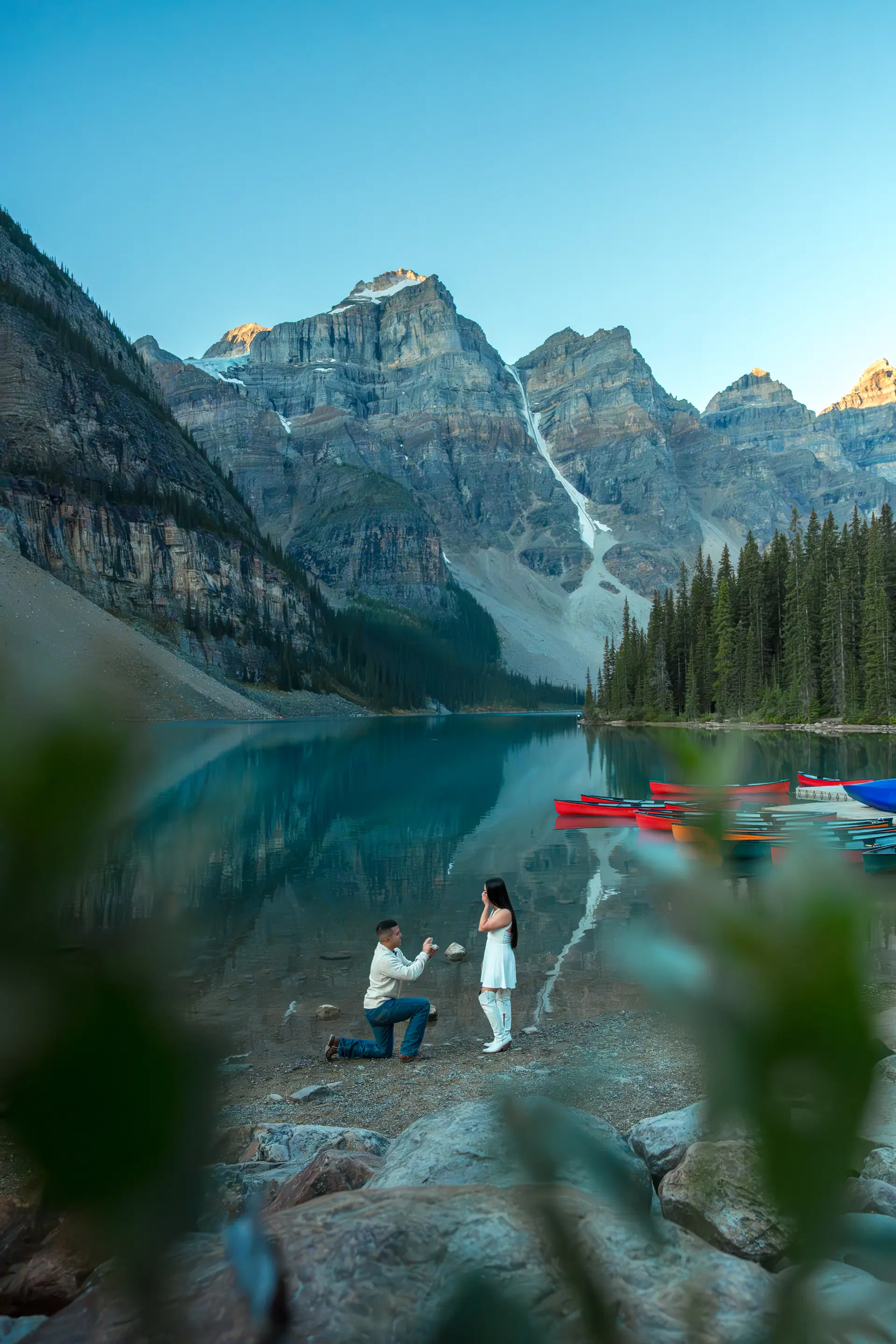 A man proposing on one knee to a woman in a white dress on the rocky shore of Moraine Lake at golden hour.