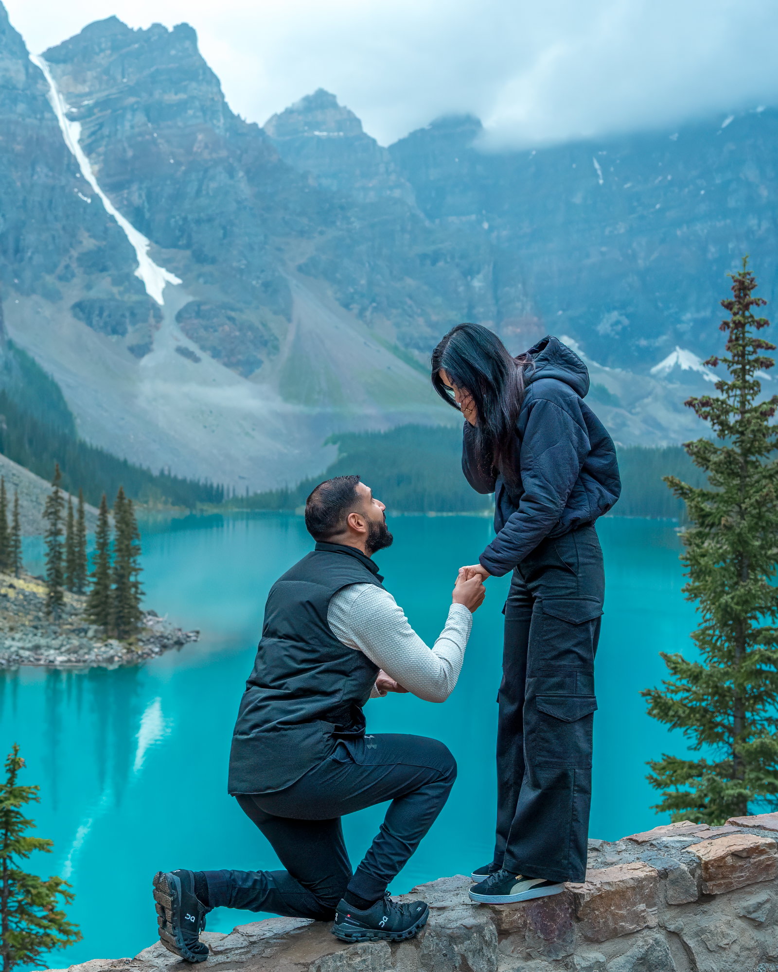 A man proposing on one knee to a woman on a rocky ledge overlooking Moraine Lake and the Valley of the Ten Peaks.